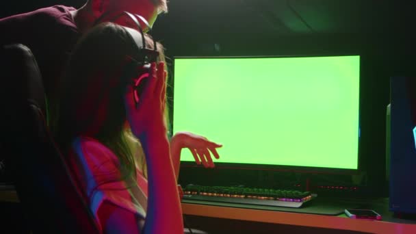 La chica apunta con la mano a la pantalla verde de la computadora en un cuarto oscuro con auriculares — Vídeos de Stock