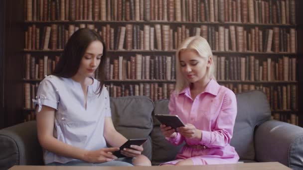 Две девушки сидят в библиотеке и читают книгу в электронном виде — стоковое видео
