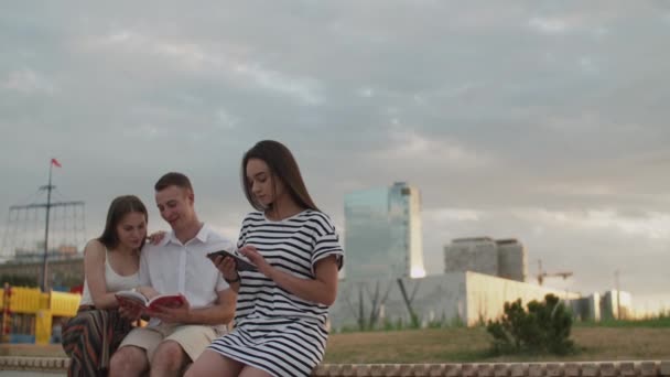 Två tjejer och en kille som sitter i parken under dagen och läser en e-bok — Stockvideo