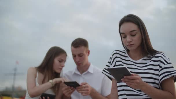 Två tjejer och en kille som sitter i parken under dagen och läser en e-bok — Stockvideo