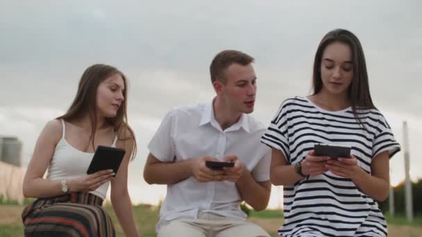 Två tjejer och en kille som sitter i parken under dagen och läser en e-bok — Stockvideo