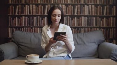 Bir kız kütüphanede bir fincan kahve içip e-kitap okuyor.