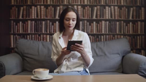 A girl drinks a cup of coffee in the library and reads an e-book — Stock Video
