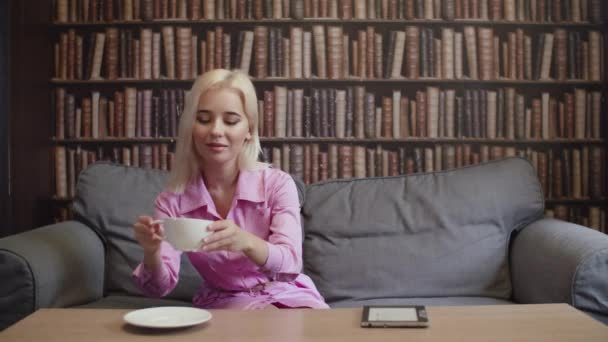 Дівчина п'є чашку кави в бібліотеці і читає електронну книгу — стокове відео