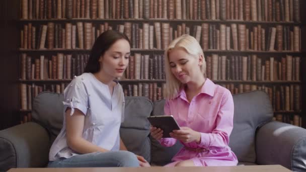 Две девушки сидят в библиотеке и читают книгу в электронном виде — стоковое видео