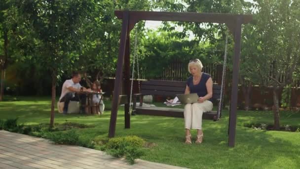 An elderly woman is sitting on a wooden swing in summer, holding a computer in her hands, filling out documents — Stock Video