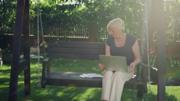 An elderly woman is sitting on a wooden swing in summer, holding a computer in her hands, filling out documents — Stock Video