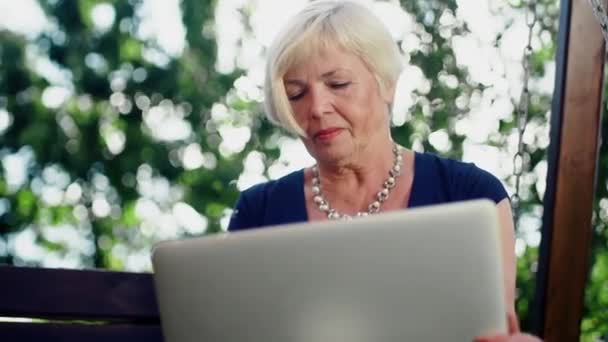 Close-up van een oudere vrouw die in de zomer op een houten schommel zit, met een computer in haar handen, documenten invullen — Stockvideo
