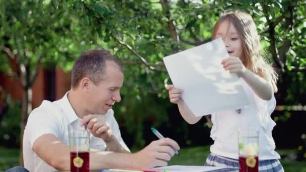 Um homem desenha com uma menina em uma mesa fora, no verão em um dia ensolarado quente — Vídeo de Stock