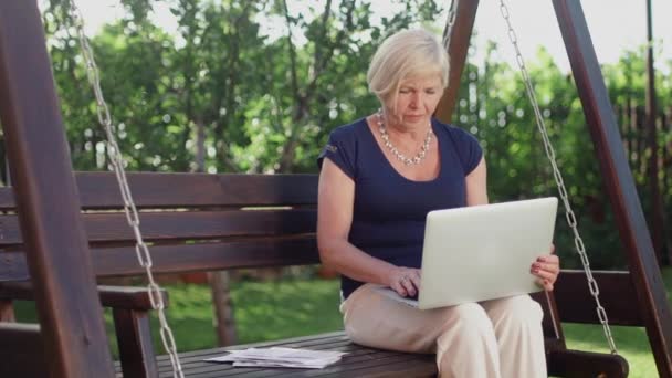 Close-up de uma mulher idosa sentada em um balanço de madeira no verão, segurando um computador em suas mãos, preenchendo documentos — Vídeo de Stock