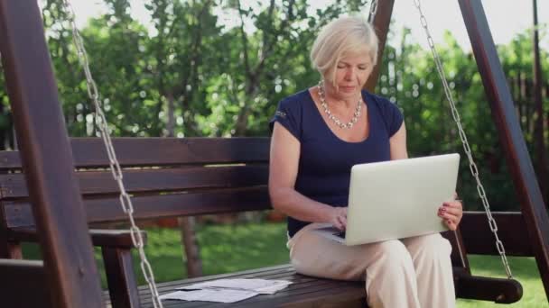 Close-up van een oudere vrouw die in de zomer op een houten schommel zit, met een computer in haar handen, documenten invullen — Stockvideo