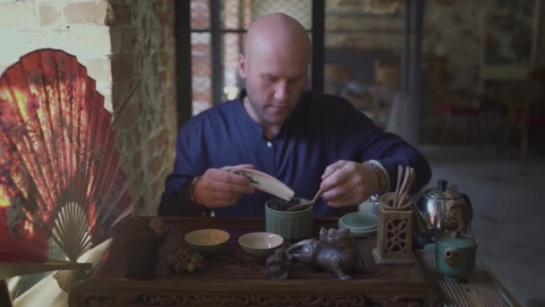 En ung man i en tebutik lägger teblad i en träsked med en spatel av trä — Stockvideo