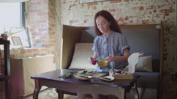 Het meisje zit aan tafel en schenkt groene thee in een theekopje en proeft het. — Stockvideo