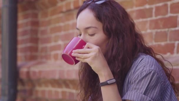 Uma linda garota de cabelos encaracolados bebe chá e desfruta de seu sabor na rua — Vídeo de Stock