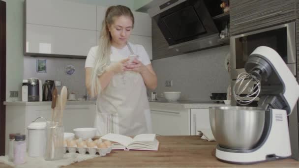 Uma menina na cozinha está procurando receitas na Internet em seu telefone — Vídeo de Stock