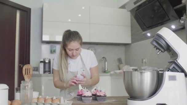 Une belle fille dans la cuisine prend des photos de cupcakes préparés sur son téléphone. Travail à la main — Video