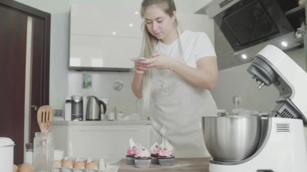 Uma linda garota na cozinha tira fotos de cupcakes preparados em seu telefone. Trabalho artesanal — Vídeo de Stock