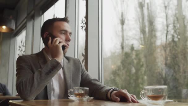 Un homme en costume de cérémonie assis à une table de restaurant avec une tasse de thé, parlant sur un téléphone portable — Video