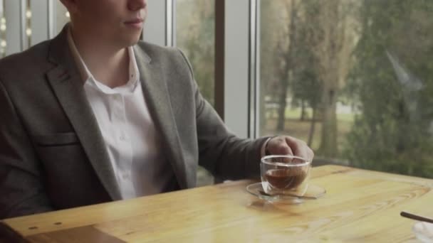 Un hombre de negocios se sienta cerca de la ventana, levanta un vaso de té verde y lo bebe. — Vídeo de stock