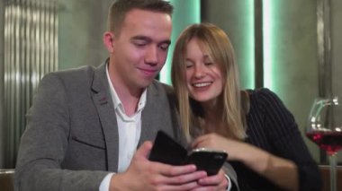 Çekici bir çift akıllı telefon kullanarak masada oturuyor, internette geziniyor, online alışverişleri tartışıyor, romantik bir akşam yemeği için restorandan yemek sipariş ediyor.