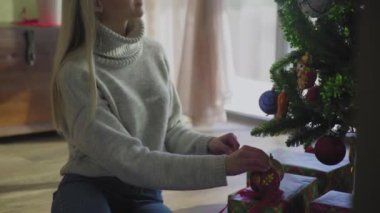 Süveter giyen genç ve güzel bir sarışın Noel ağacının yanında oturuyor ve bir Noel balosu düzenliyor. Evin penceresinin yanında altında hediyeler olan bir Noel ağacı..