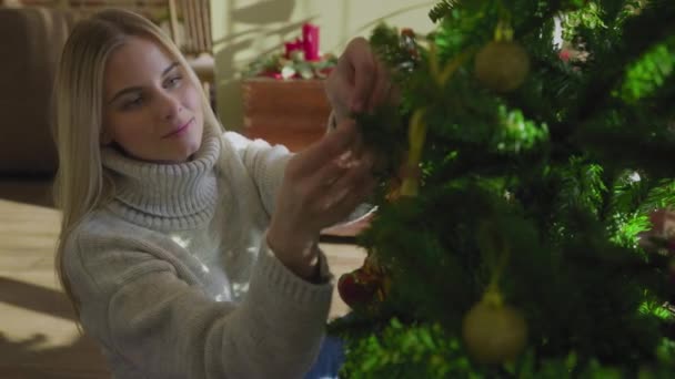 En vacker ung blondin i en tröja sitter bredvid julgranen och sätter på sig en julboll. En julgran vid fönstret i huset med en massa presenter under. — Stockvideo