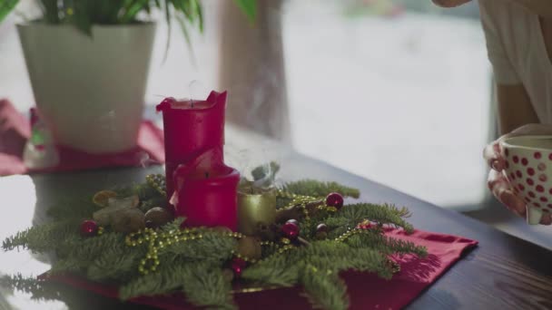An unrecognizable girl blows out an evergreen wreath with two red candles and one golden candle. Christmas traditions and customs of Christians on the eve of the holiday. A symbol of the holiday. — Stock Video
