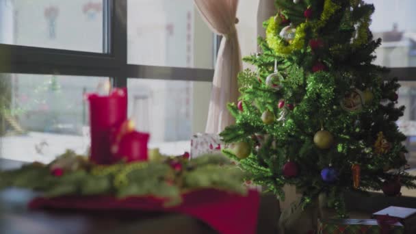 Ένα αειθαλές στεφάνι με δύο κόκκινα κεριά και ένα χρυσό κερί. Στο βάθος, ένα στολισμένο χριστουγεννιάτικο δέντρο. Χριστουγεννιάτικες παραδόσεις και έθιμα των Χριστιανών την παραμονή των εορτών. — Αρχείο Βίντεο