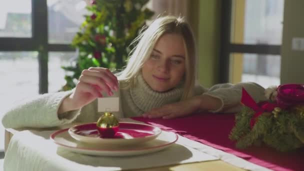 Una joven hermosa mujer se apoya sobre la mesa con las manos, mirando una bola de Año Nuevo. Preparándose para la Navidad. Humor festivo. — Vídeo de stock