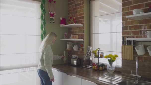 Μια νεαρή όμορφη ξανθιά έρχεται στην κουζίνα και διορθώνει αντικείμενα και διάφορα διακοσμητικά. Χριστούγεννα — Αρχείο Βίντεο