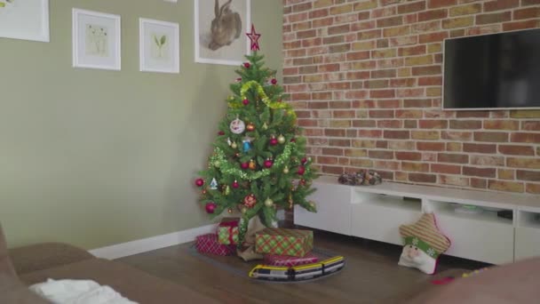 No canto da casa há uma árvore de Natal com um monte de presentes sob ele, e um trem de brinquedo anda ao redor da árvore. Atmosfera acolhedora — Vídeo de Stock