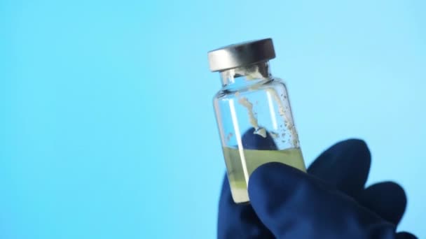 Zbliżenie dłoni w rękawiczkach lateksowych trzymających szklaną ampułkę szczepionki na niebieskim tle — Wideo stockowe