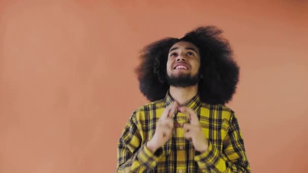 Ein junger Mann mit afrikanischer Frisur auf orangefarbenem Hintergrund drückt ihm die Daumen und betet. Emotionen auf farbigem Hintergrund. — Stockvideo