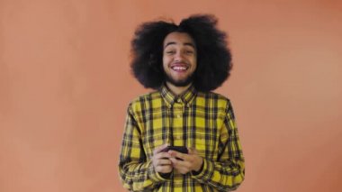 Turuncu arka planda Afrikalı saç stili olan genç bir adam telefona bakar ve mutlu olur. Renkli bir arkaplanda duygular