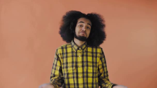 En ung mand med afrikansk frisure på orange baggrund trækker på skuldrene. Følelser på en farvet baggrund. – Stock-video