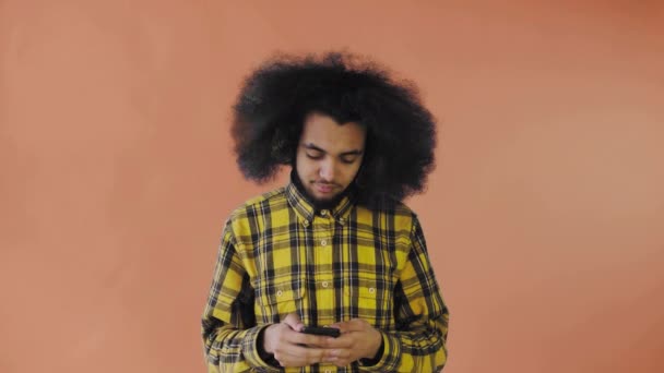 En ung man med en afrikansk frisyr på orange bakgrund pratar i sin telefon.. — Stockvideo