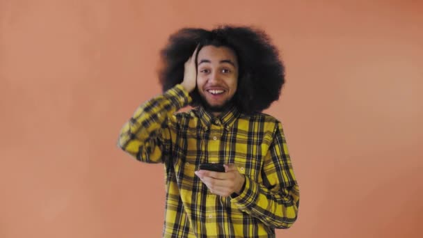 Um jovem com um penteado africano em um fundo laranja olha para o telefone e fica feliz surpreso. Emoções em um fundo colorido — Vídeo de Stock