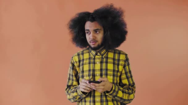 Un giovane con un'acconciatura africana su uno sfondo arancione guarda il telefono e dubita. Emozioni su sfondo colorato — Video Stock