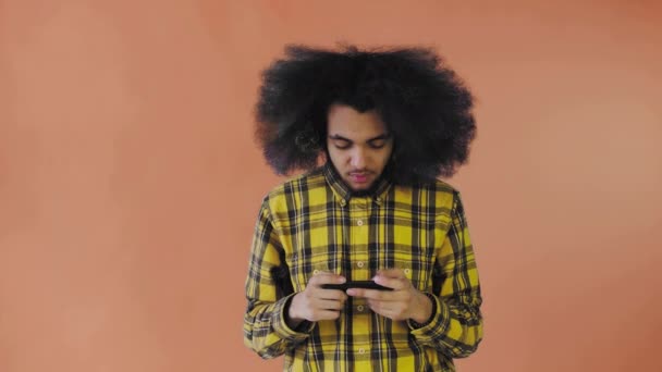 一个有着橙色背景的非洲发型的年轻人在电话里玩。在彩色背景上 — 图库视频影像