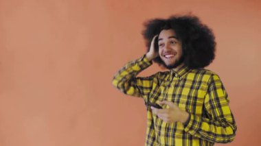 Turuncu arka planda Afrikalı saç stili olan genç bir adam telefona bakar ve mutlu bir şekilde şaşırır. Renkli bir arkaplanda duygular