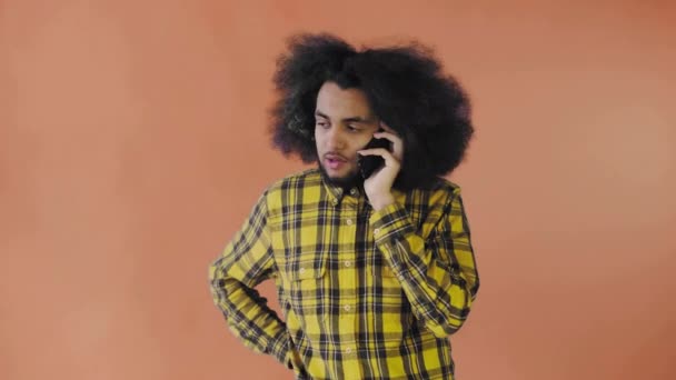 Um jovem com um penteado africano num fundo laranja está a falar ao telefone. Em um fundo colorido — Vídeo de Stock