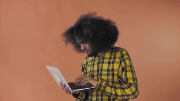 A young man with an African hairstyle on an orange background is typing on a laptop. On a colored background — Stock Video