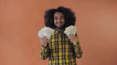 Elinde banknot yelpazesi olan mutlu bir Afro-Amerikalı adam, turuncu bir arka planda izole bir şekilde kameraya bakıyor.