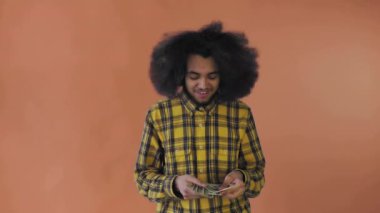 Elinde banknotlar tutan mutlu bir Afro-Amerikalı adam, onları sayıyor ve kameraya bakıyor, turuncu bir arka planda tek başına duruyor.