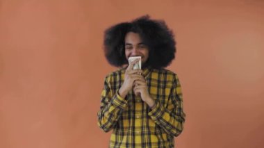 Elinde banknotlar tutan, onları öpen ve kameraya bakan mutlu bir Afro-Amerikalı adam turuncu bir arka planda tek başına duruyor.