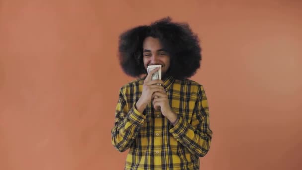 Щасливий афро-американець, який тримає в руках банкноти, цілує їх і дивиться на камеру, стоїть ізольований на помаранчевому фоні. — стокове відео