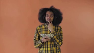 Elinde para tutan ve neye harcayacağını düşünen mutlu bir Afro-Amerikalı turuncu bir arka planda tek başına duruyor.