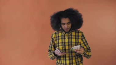 Elinde banknotlar tutan mutlu bir Afro-Amerikalı adam, onları sayıyor ve kameraya bakıyor, turuncu bir arka planda tek başına duruyor.