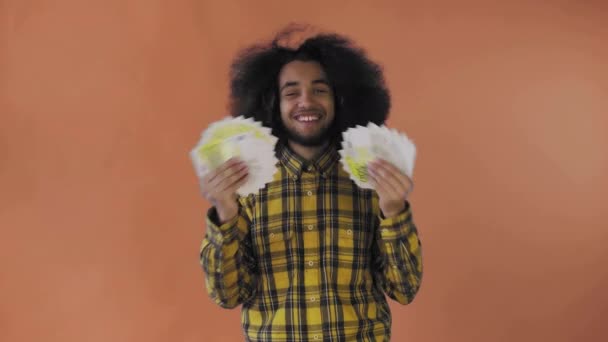 Un hombre afroamericano feliz sosteniendo un abanico de billetes en sus manos y mirando a la cámara, parado aislado sobre un fondo naranja — Vídeo de stock