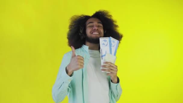 Счастливый афроамериканец смотрит в камеру, держит в руке туристические ваучеры, стоит изолированный на желтом фоне — стоковое видео